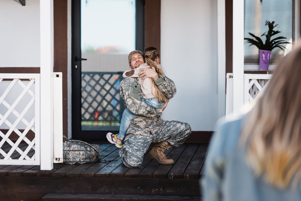 Sorridente militare uomo abbracciare figlia, mentre seduto in ginocchio sulla soglia con la donna sfocata in primo piano - Foto, immagini