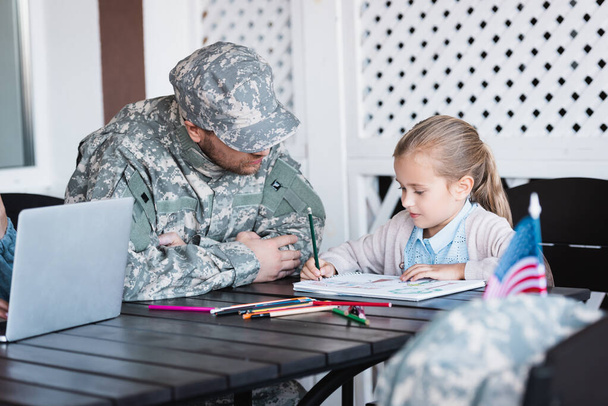 Ojciec w mundurze wojskowym siedzi obok córki rysując kolorowymi ołówkami w domu na niewyraźnym pierwszym planie  - Zdjęcie, obraz