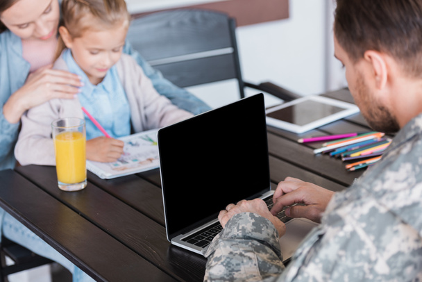 Man in military uniform typing on laptop with blank screen, while sitting at table with blurred woman and girl on background - Fotografie, Obrázek