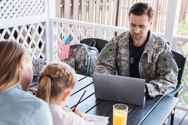 Military serviceman with necklace using laptop, while sitting at table with blurred woman and daughter on foreground - Zdjęcie, obraz
