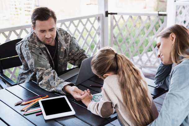 Padre en uniforme militar mirando dibujo hija, mientras está sentado en la mesa con el ordenador portátil y la tableta digital - Foto, Imagen