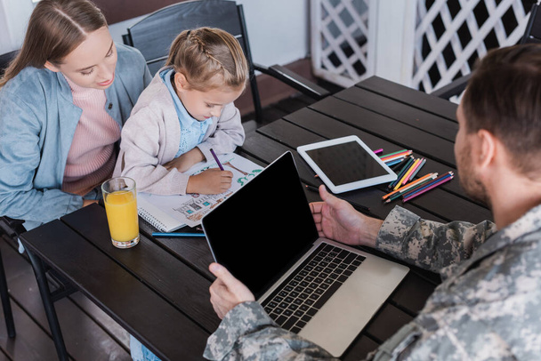 Militar serviceman usando el ordenador portátil y mirando a la chica sentada con la mujer y el dibujo con lápices de colores en la mesa - Foto, Imagen