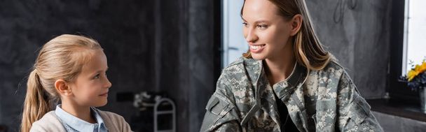 Happy woman in military uniform and girl looking at each other, while sitting in kitchen, banner - Photo, Image