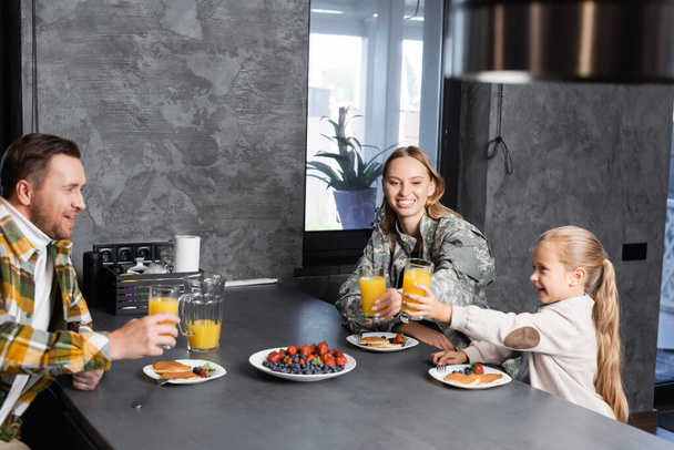 Happy family toasting with glasses of juice, while sitting at table with fruits and pancakes in kitchen - Photo, Image