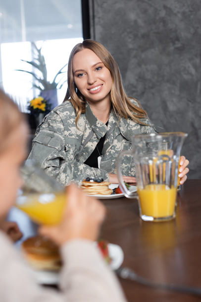 Glimlachende vrouw met blond haar aan tafel met pannenkoeken en kruik sap in de keuken met wazig meisje op de voorgrond - Foto, afbeelding