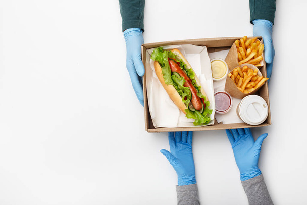 Υγιεινό μενού στο εστιατόριο fast food. Courier σε λαστιχένια γάντια δίνει κουτί με χοτ ντογκ, τηγανητές πατάτες, κέτσαπ και σάλτσα, takeaway καφέ, στον πελάτη - Φωτογραφία, εικόνα