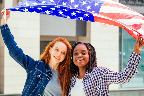 lesbian redhaired ginger woman and her african american wife holding USA flag in downtown street - Foto, immagini