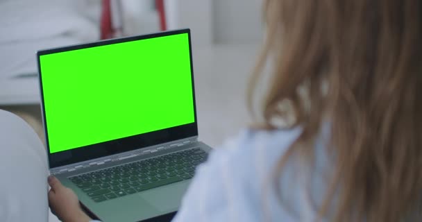 Νεαρή γυναίκα στο σπίτι εργάζεται σε έναν φορητό υπολογιστή με πράσινη οθόνη mock-up. Κάθεται σε έναν καναπέ στο άνετο σαλόνι του. Πάνω από τη λήψη της κάμερας ώμου - Πλάνα, βίντεο