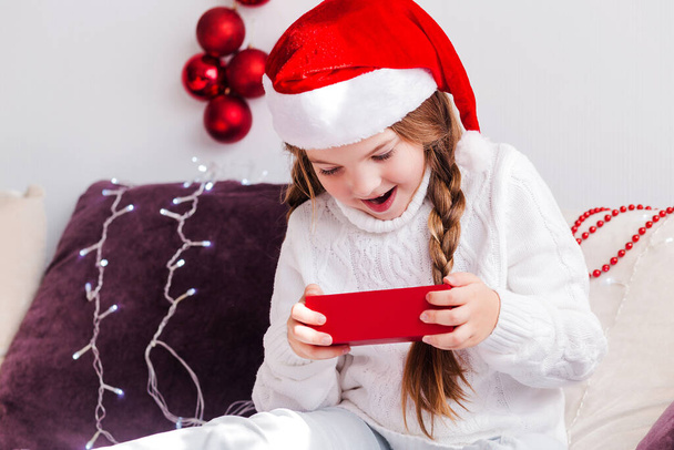 Das kleine Mädchen in weißem Pullover und Nikolausmütze betrachtet ein Geschenk in einer roten Schachtel und ist überrascht. Horizontales Foto - Foto, Bild