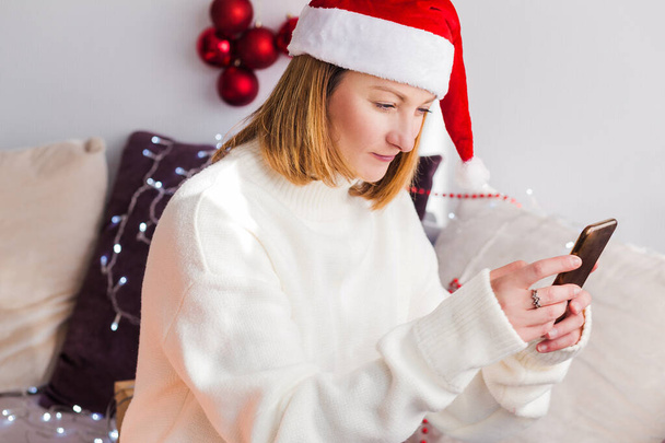 A young woman in a white sweater, Santa Claus hat sits at home on the couch with a mobile phone in her hands. Woman looking at cell phone. Horizontal photo - Foto, Imagen
