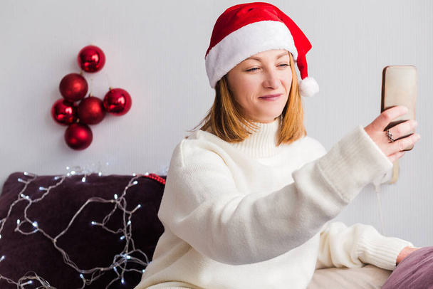 Młoda kobieta w białym swetrze, kapelusz Świętego Mikołaja siedzi w domu na kanapie z telefonem komórkowym w rękach. Kobieta patrzy na komórkę i się uśmiecha. Zdjęcie poziome - Zdjęcie, obraz