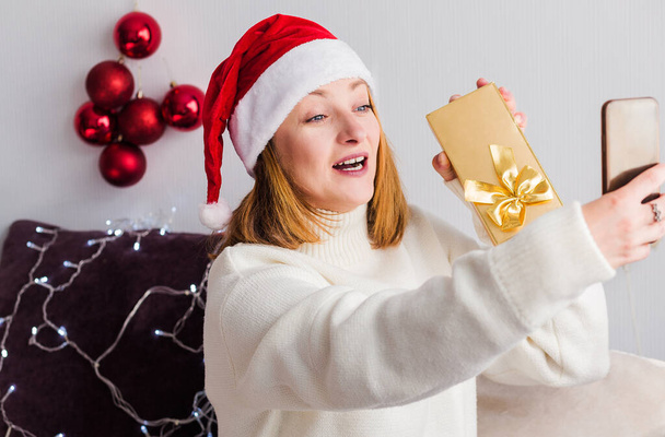 Mladá žena v bílém svetru, Santa Claus klobouk sedí doma na gauči s mobilním telefonem v ruce. Žena mluví na videohovoru, usmívá se a drží v rukou dárek. Vodorovná fotografie - Fotografie, Obrázek