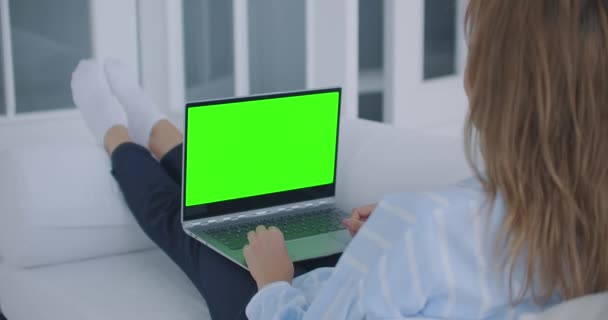 Een jonge vrouw zit met een laptop op schoot met een groen scherm tijdens de quarantaine. chromakey op het laptopscherm. Maak een videoconferentie en praat met het groene scherm - Video