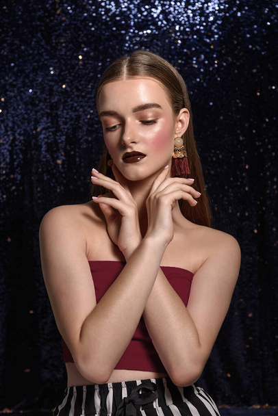 Fashionable girl with bright, professional evening make-up. Burgundy lipstick, clean, natural face, French manicure. Hair is parted, large hanging gold earrings with pearls. Shiny  blue background - Valokuva, kuva