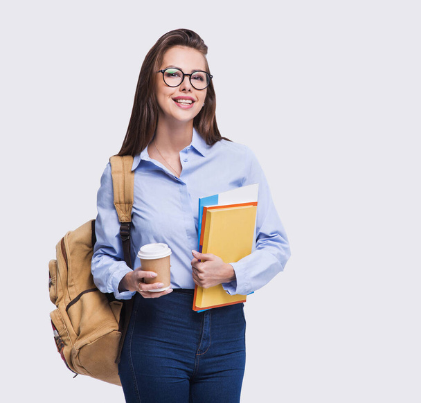 Studio portrait of a beautiful student girl. Young smiling student woman holding books and coffee cup. Isolated on grey background - Фото, изображение