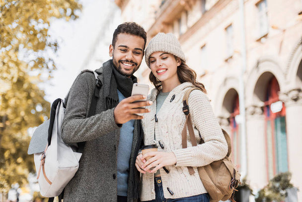 Beautiful happy couple using smartphone. Young joyful smiling woman and man looking at mobile phone in a city in autumn. Technology, travel, tourism, students concept - Fotó, kép