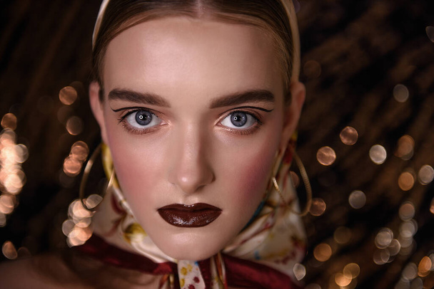 Close-up portrait of a girl with evening, fashion make-up, maroon lipstick, clear skin, pink rumanian, round gold earrings in her ears, head covered with a fashionable satin headscarf. Golden glitter  - 写真・画像