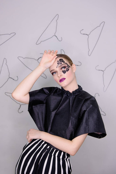 Fashion portrait of a girl with clean skin, bright professional make-up, purple lipstick, face with black ivy tattoo around her eyes with rhinestones. Gray background, pattern of hangers. smoky eyes - 写真・画像