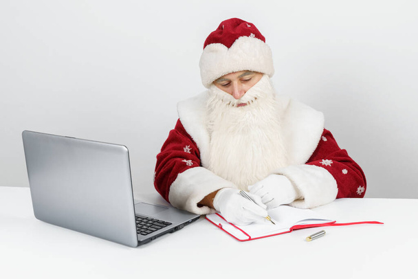 Weihnachten und Neujahr. Der Weihnachtsmann sitzt an seinem Schreibtisch und schreibt in ein Notizbuch. - Foto, Bild