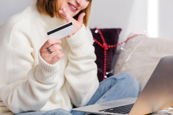Młoda kobieta w białym swetrze, kapelusz Świętego Mikołaja dokonuje zakupów online przy użyciu notesu i karty kredytowej. Noworoczne zakupy i świąteczna wyprzedaż. Zdjęcie poziome - Zdjęcie, obraz