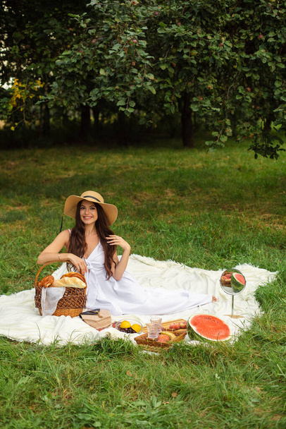 Πορτρέτο ενός νεαρού όμορφου κοριτσιού με ακόμα και λευκά δόντια, ένα όμορφο χαμόγελο σε ψάθινο καπέλο και μακρύ λευκό φόρεμα έχουν ένα πικνίκ στον κήπο - Φωτογραφία, εικόνα