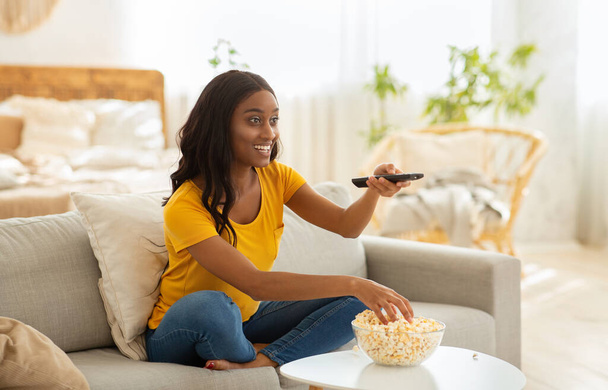 Молодая афроамериканка сидит на диване, ест попкорн и смотрит интересное шоу по телевизору, в помещении - Фото, изображение