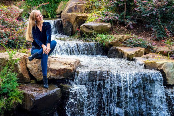Una hermosa modelo rubia se sienta cerca de una cascada en un parque local - Foto, imagen