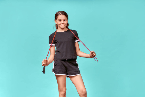 Schattig tiener meisje in zwart sportkleding met springtouw kijken naar de camera en glimlachen tijdens het hebben van training, geïsoleerd staan over blauwe achtergrond - Foto, afbeelding