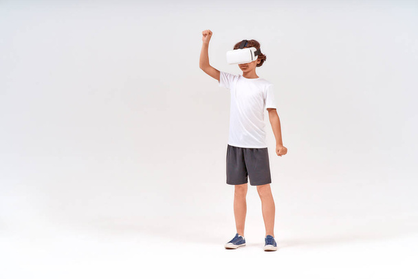 Full-length shot of teenage boy in sport clothes wearing virtual reality or 3d glasses exercising isolated over grey background, studio shot - Foto, Bild