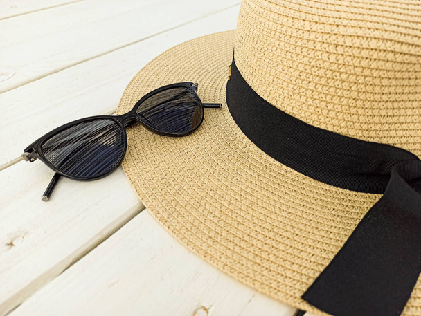 黒いサングラスで白い木製の背景に黒いリボンと美しい女性のわらの帽子。テーブルの上には眼鏡の帽子がかかっている。夏の頭飾りとサングラス. - 写真・画像