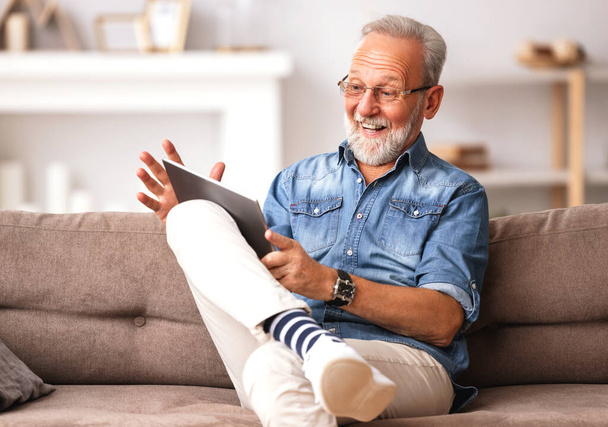 Szczęśliwy starszy brodaty mężczyzna w ubraniach casual przeglądania tabletek i uśmiecha się siedząc na kanapie w hom - Zdjęcie, obraz