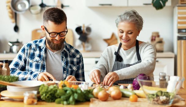 Feliz familia hombre barbudo positivo con alegre madre madura cortando verduras frescas mientras preparan ensalada saludable juntos en la acogedora cocina en hom - Foto, imagen