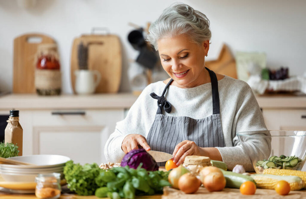 Alegre ama de casa de mediana edad en delantal sonriendo mientras prepara un plato saludable con verduras frescas en la cocina casera - Foto, imagen