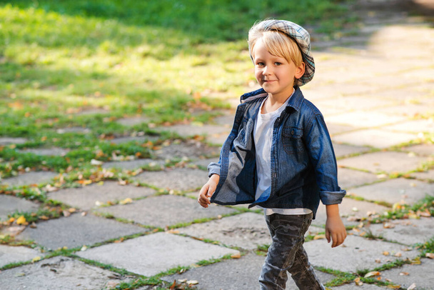 Happy child walking in the park. Stylish kid posing outdoors. Cute little boy in summer outfit. Child wearing trendy clothes and hat. Kid having fun outside in the park. - Photo, image