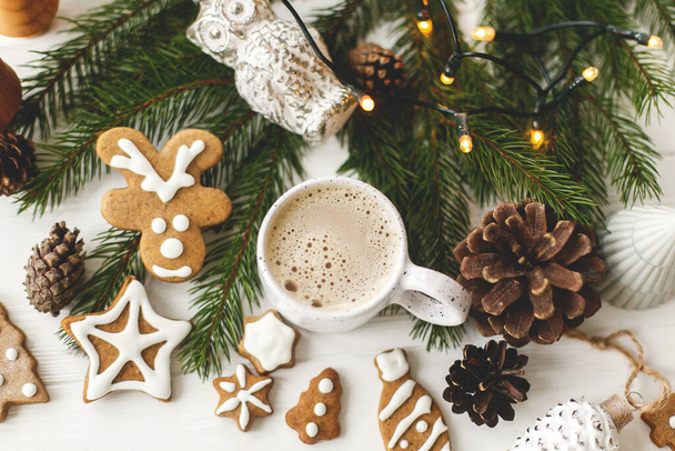 Christmas gingerbread cookies, coffee in stylish white cup, pine cones  and warm lights on white wooden table. Hello winter, cozy moody image top view - Фото, изображение