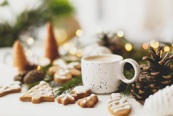 Christmas gingerbread cookies, coffee in stylish white cup, pine cones  and warm lights on white wooden table. Hello winter, cozy moody image with selective focus - Foto, imagen