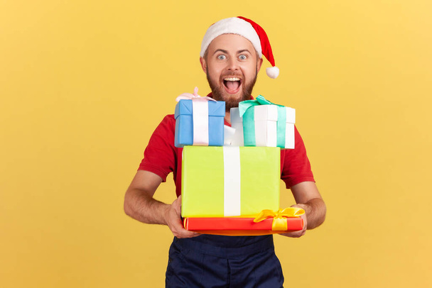 Extremely happy courier in blue uniform and red santa claus hat holding and showing christmas gifts looking at camera with toothy smile. Indoor studio shot isolated on yellow background - Фото, зображення