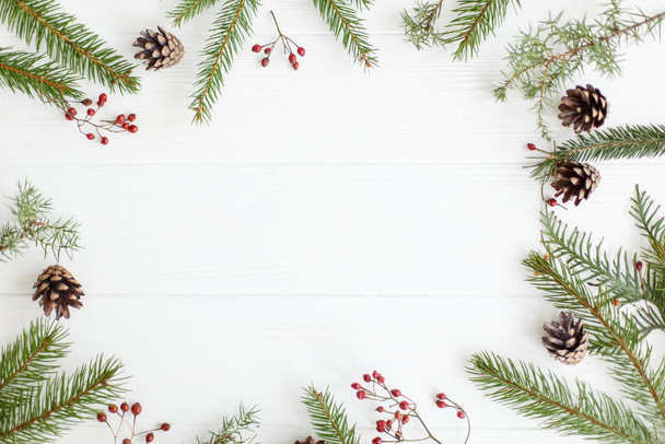 Рождественские елки, красные ягоды и сосновые шишки рамка на белом дереве, минималистская плоская укладка. Сезонная открытка макетирует место для текста. Счастливого Рождества и счастливых праздников!! - Фото, изображение