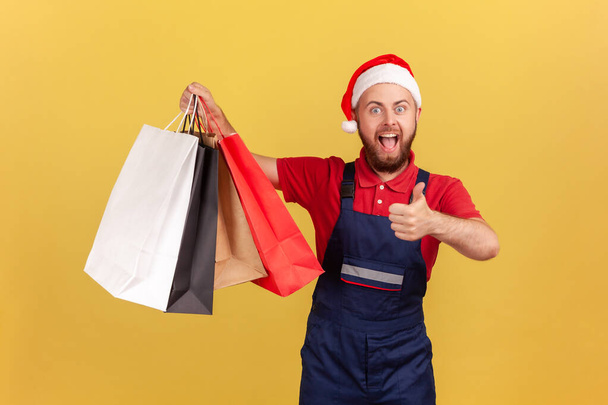 Happy excited male courier in uniform and santa claus hat showing thumbs up holding paper shopping bags, likes express delivery. Indoor studio shot isolated on yellow background - Foto, imagen