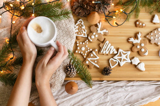 Hände mit warmem Kaffee auf dem Hintergrund weihnachtlicher Lebkuchen, kuscheliger Strickpullover, Tannenzweige mit Tannenzapfen und Lichtern. Hallo Winter. Frohe Weihnachten und frohe Feiertage! - Foto, Bild