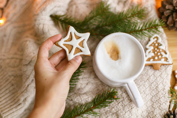 居心地の良いニットセーター、コーヒー、モミの枝や暖かい光を背景にクリスマススタージンジャーブレッドクッキーを保持手。冬だ。メリークリスマスとハッピーホリデー! - 写真・画像