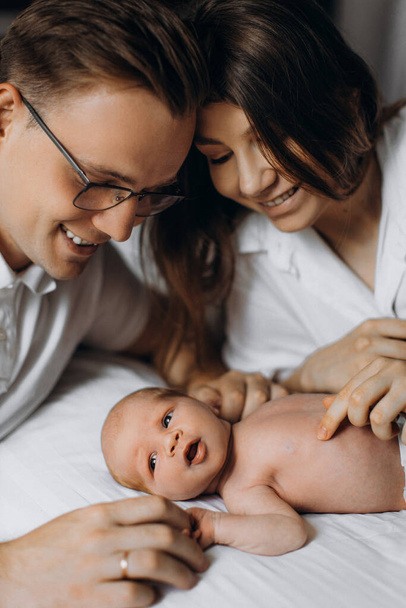 Adorable newborn baby laying at the bed with caring parents, loving dad gently hold arms of little daughter, lovely mom smiling, enjoy tender moments, young family concept - Foto, Imagem