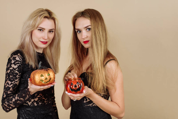 Dos jóvenes mujeres sonrientes sostienen calabazas de Halloween sobre un fondo beige. Modelos en vestidos negros en el estudio celebran el 31 de octubre. - Foto, Imagen
