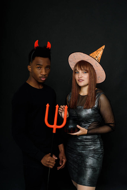 Jovem negro e mulher caucasiana celebram Halloween em estúdio em trajes em fundo preto. Sorrindo jovens amigos. - Foto, Imagem