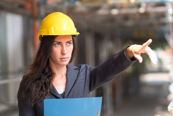 İçine çekilen kadın mühendis sarı koruyucu bir şapka takıyor ve inşaat alanını işaret ediyor. - Fotoğraf, Görsel