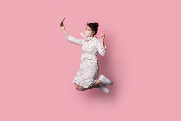 Brunette vrouw met medisch masker op het gezicht is springen in een zomer jurk en het maken van een selfie op een roze studio muur - Foto, afbeelding