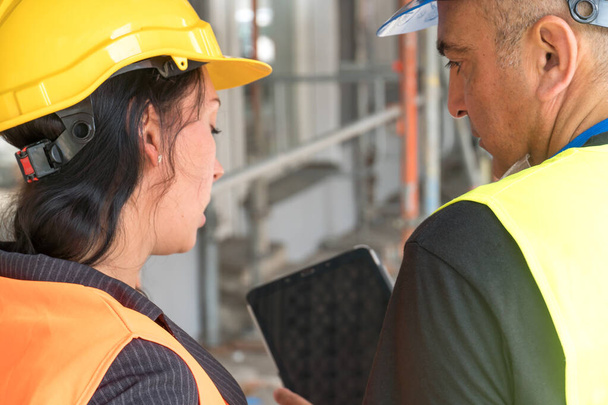 Deux travailleurs de la construction dos tourné portant des gilets de sécurité et des casques vérifiant leurs plans de travail sur une tablette numérique - Photo, image