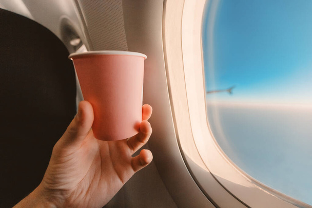 An airplane passenger's hand holds a paper Cup of tea against the blue sky in the window. Concept of the beginning of the journey and liquid consumption during the flight - Foto, Imagem