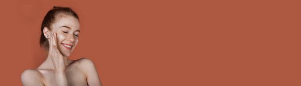 Donna caucasica con i capelli rossi e le spalle nude è in posa su un muro rosso studio con spazio libero, mentre il sorriso e l'applicazione di crema anti invecchiamento sul viso - Foto, immagini