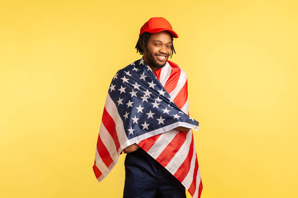 Positive afro-american man in red cap with dreadlocks standing wrapped in American flag looking at camera and smiling, proud of his country. Indoor studio shot isolated on yellow background - Фото, зображення